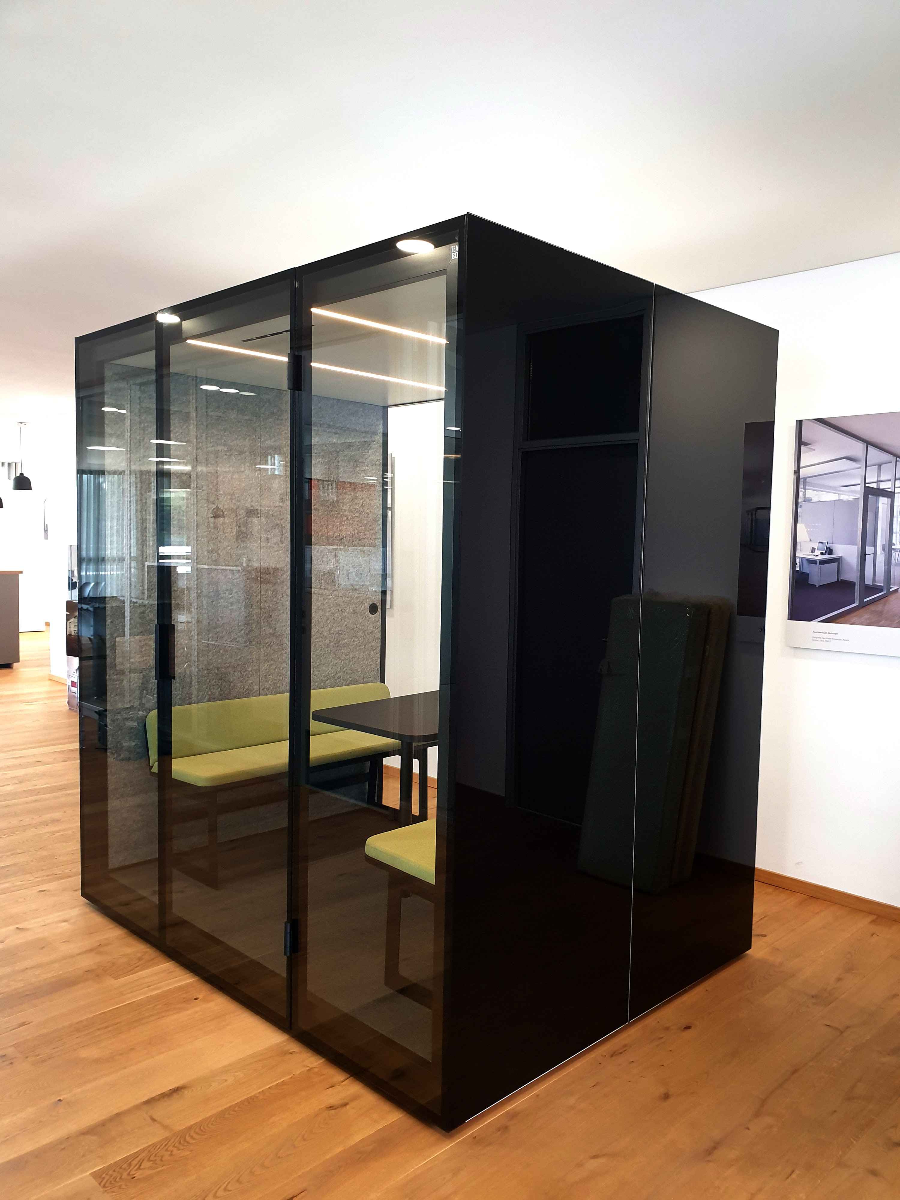 Architektonische Besprechungsbox aus dunklem Glas. Im Inneren steht ein Tisch und ein hellgrünes Sitzarrangement, ideal für Meetings.