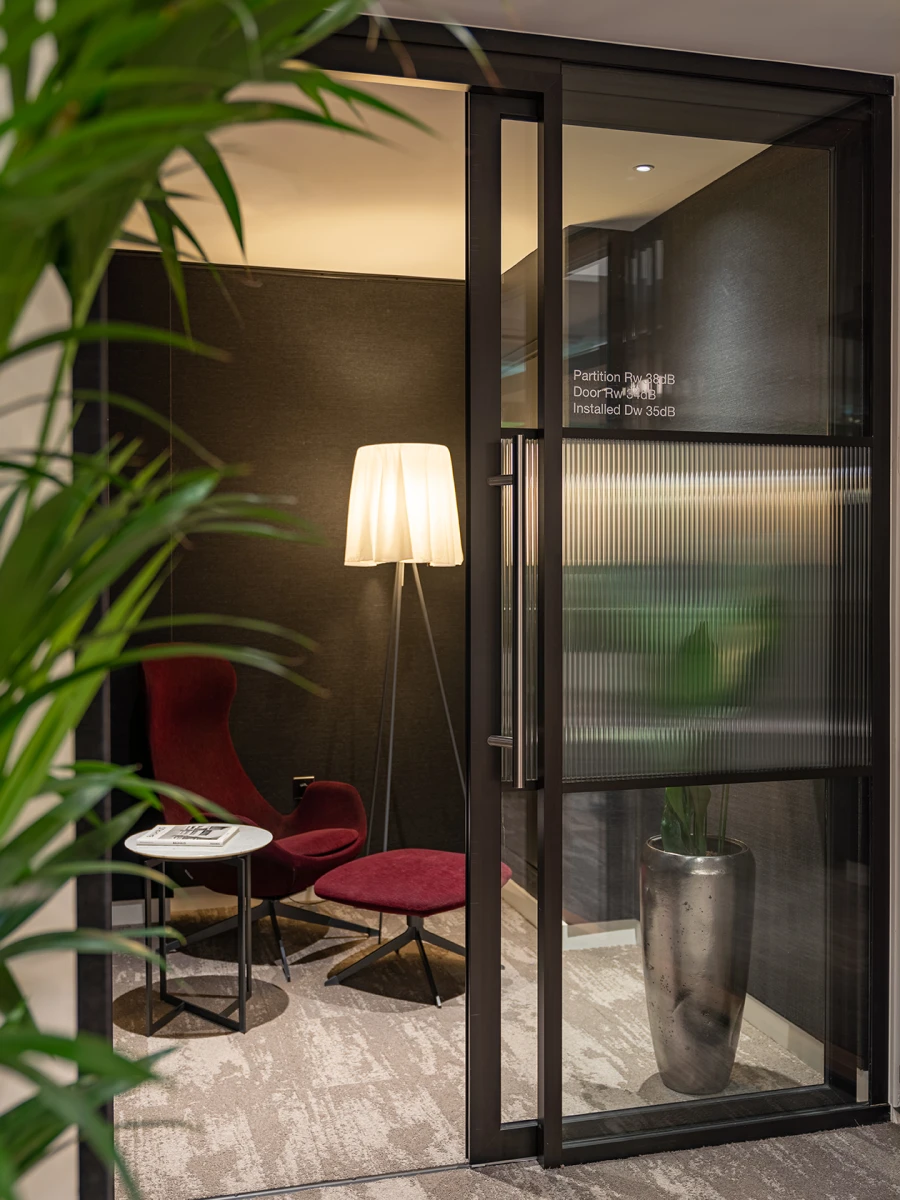 Modernes Interieur mit schwarzen Glastrennwänden und matten Verglasungen. Im Hintergrund: Rote Loungemöbel und eine Stehleuchte in einem eleganten Showroom-Ambiente.