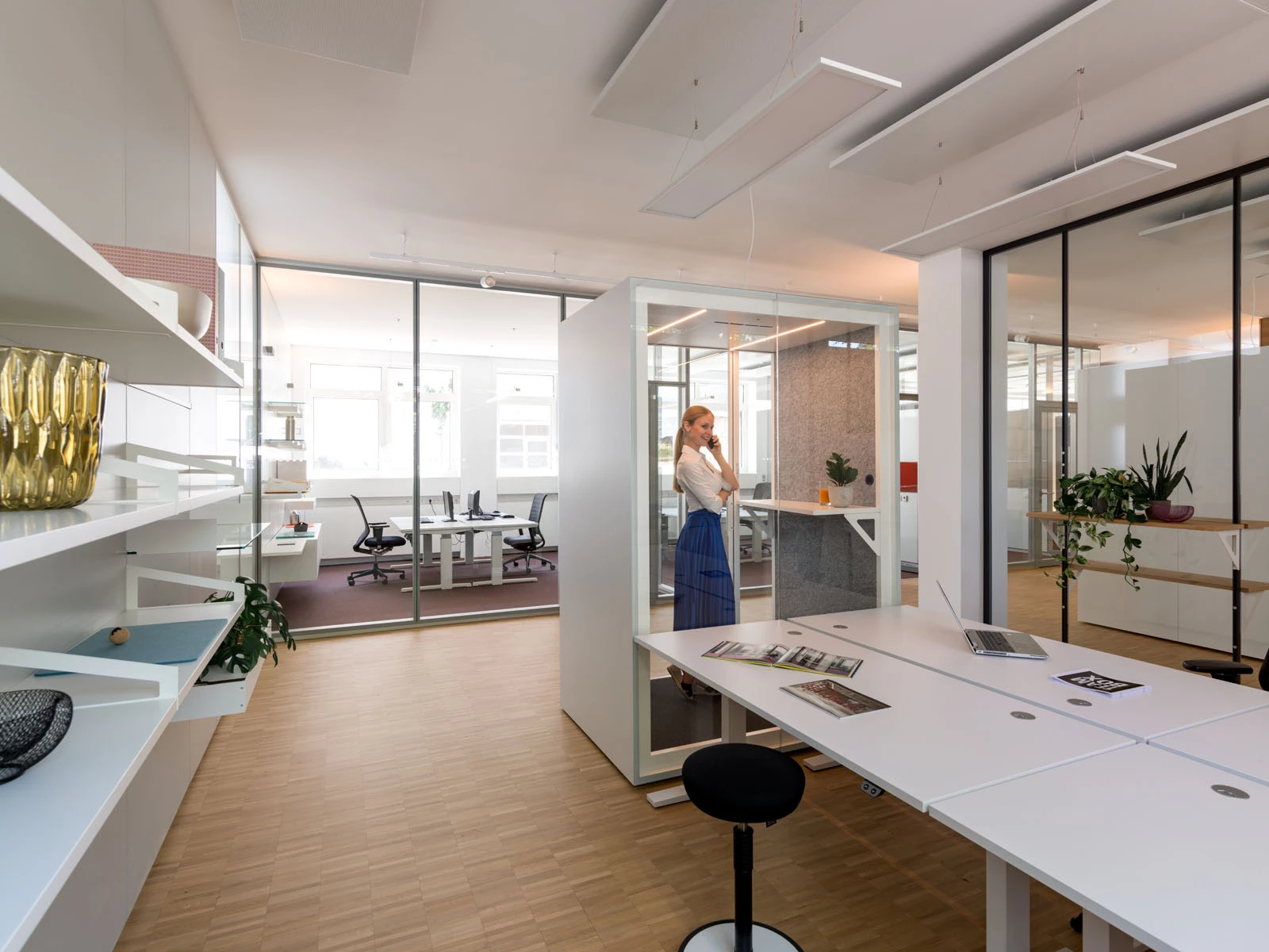 Großes, modernes Büro mit weißen Tischen, einer Akustik-Teambox und vielen Glaswänden für offene Arbeitsplätze.