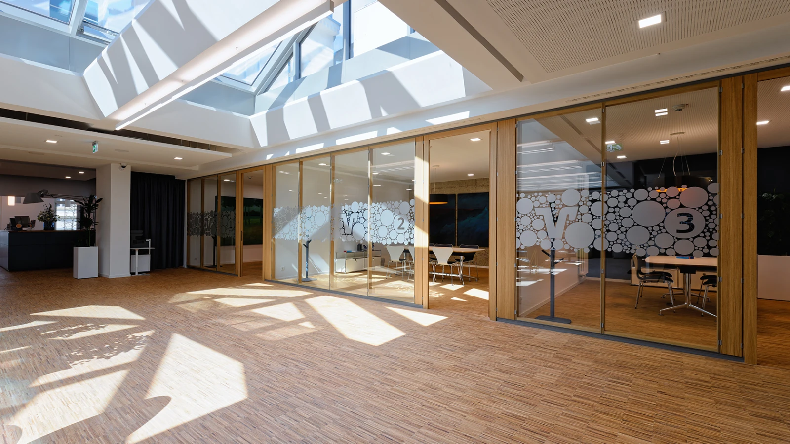 Heller, moderner Innenraum mit Holzfußboden und großen Glasscheidewänden. Die Bereiche sind mit abstrakten Kreismotiven gestaltet und von viel Tageslicht durchflutet.