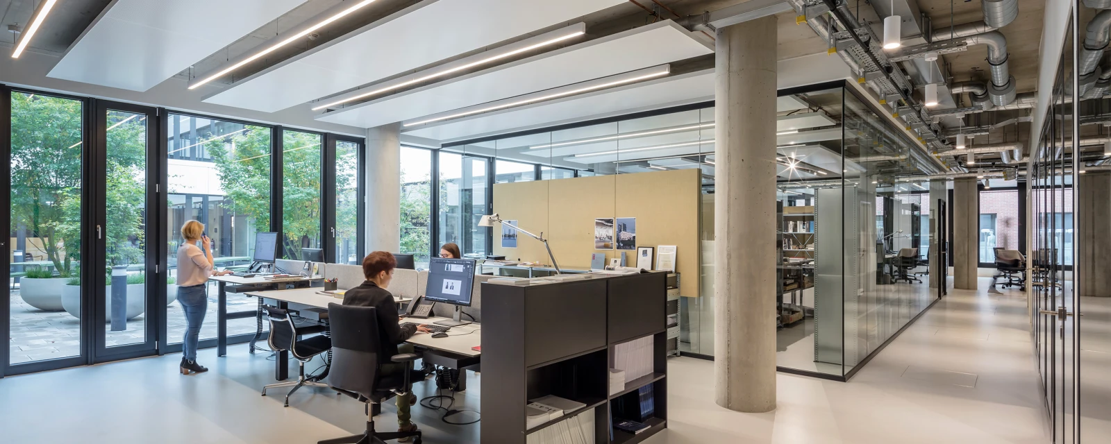 Großes, modernes Büro mit natürlichen Lichtquellen. Mitarbeiter arbeiten in einem offenen Grundriss mit Glaswänden und einem Industrie-Look.
