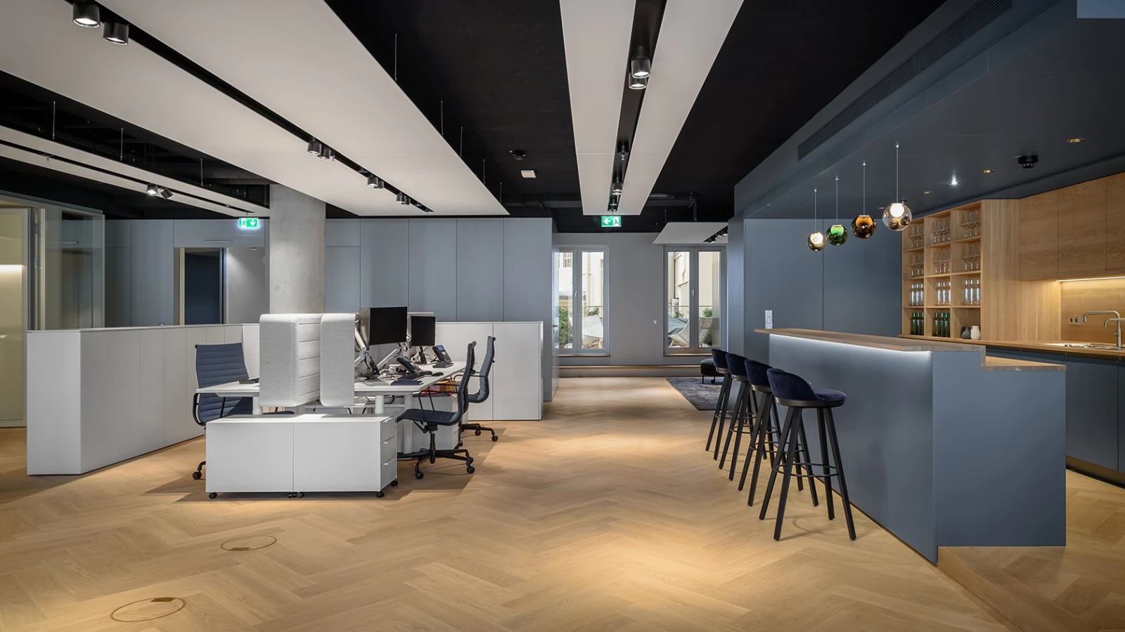 Modernes Büro- und Lounge-Ambiente: Heller Holzboden, ein Arbeitsbereich mit Schreibtischen und eine dunkle Bar mit Barhockern.