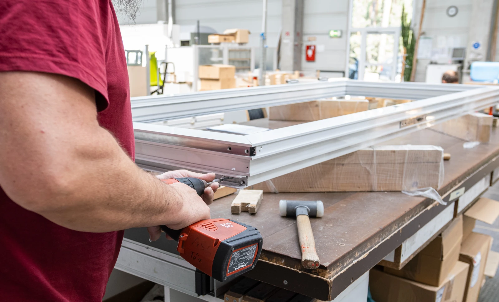 Arbeiter bearbeitet ein weißes Aluminiumrahmenprofil mit einem elektrischen Werkzeug auf einer Werkbank.