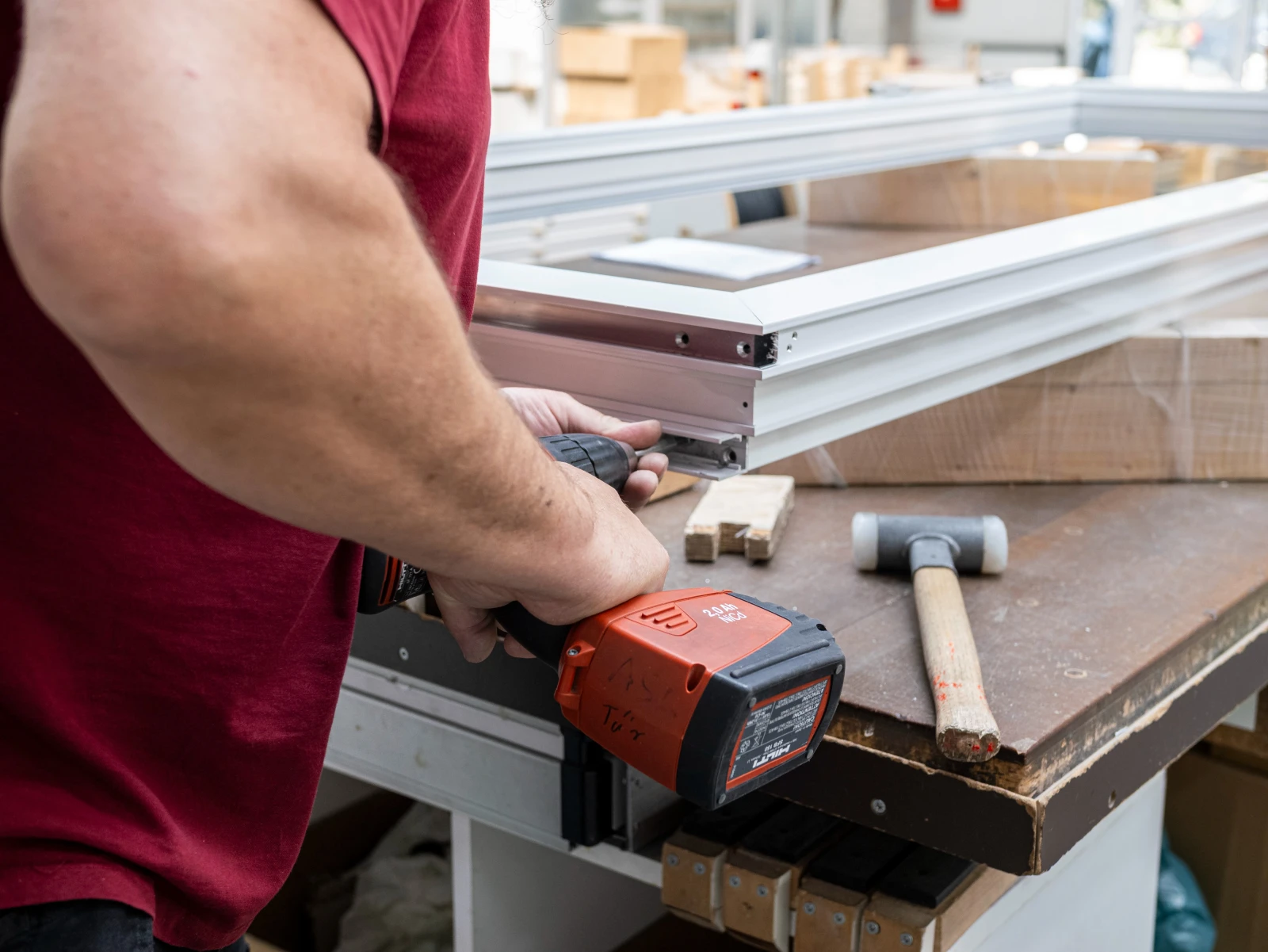 Ein Handwerker bearbeitet ein weißes Bauelement mit einem elektrischen Werkzeug. Auf dem Holztisch liegen Hammer und kleine Holzstücke.