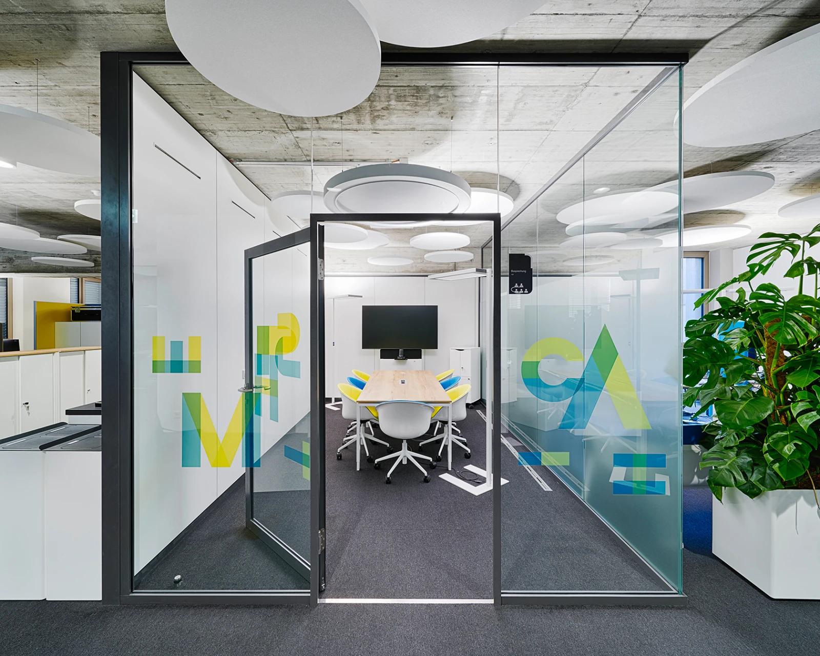 Offener Blick durch moderne Glastür in einen hellen, farblich akzentuierten Besprechungs- und Arbeitsbereich.