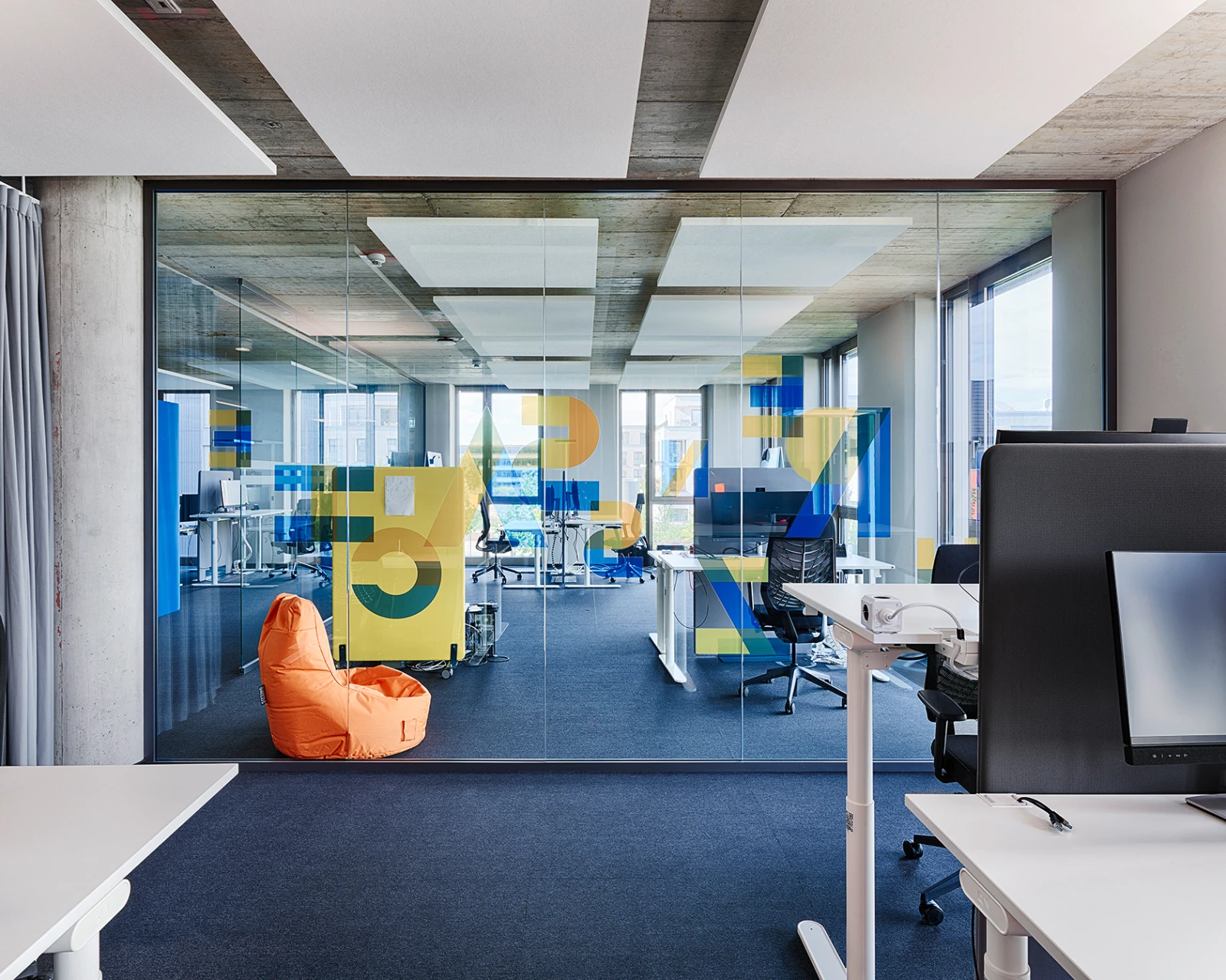 Moderner Großraumbüro-Einblick durch eine Glaswand. Sichtbares Open-Plan-Office mit Arbeitsplätzen und orangefarbenen Sitzgelegenheiten im Hintergrund.