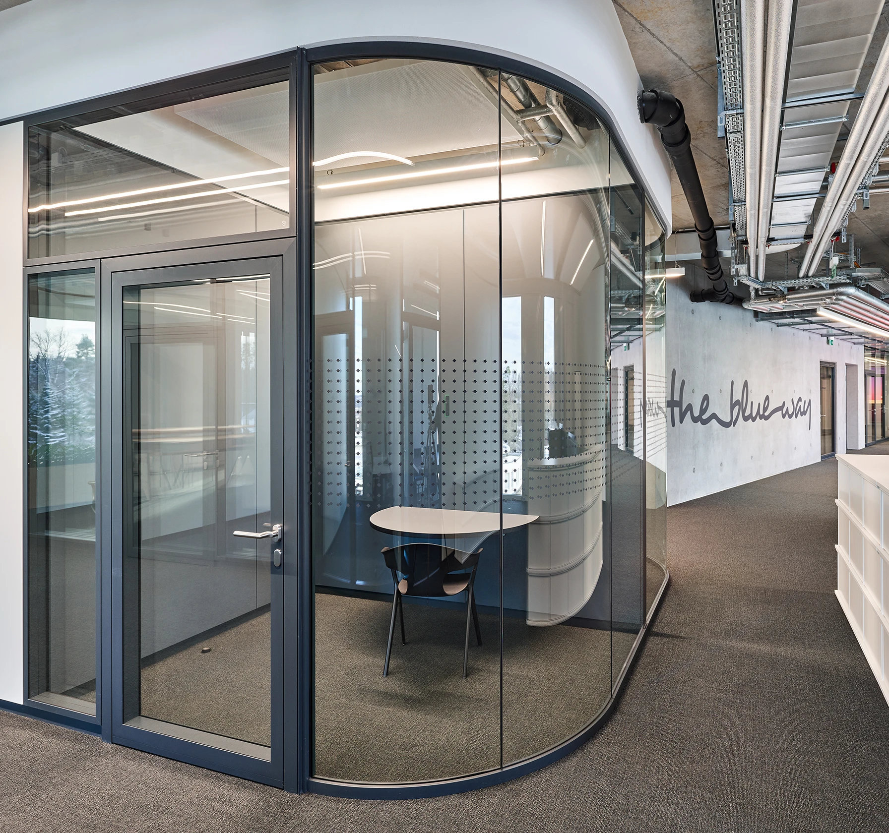 Halbrunder Besprechungsraum in einem modernen Büro, abgetrennt durch große Glaswände. Im Inneren steht ein weißer Tisch; die Rückwand zeigt perforierte Elemente und die Aufschrift „the blue way“.