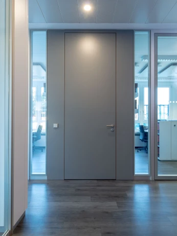 Minimalistische graue Eingangstür zwischen vertikalen Glaspartitionen in einem modernen, hellen Büroraum.