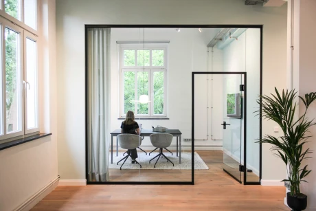 Modernes Bürointerieur: Glastrennwände trennen einen hellen Arbeitsbereich mit Schreibtisch und großen Fenstern.