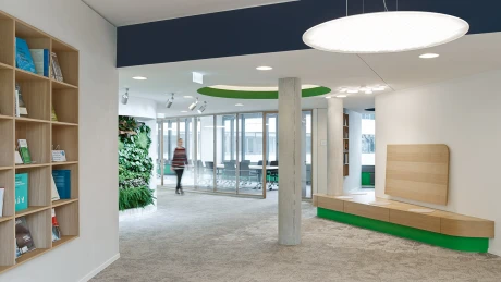 Modernes Bürointerieur mit einem großen runden Leuchtkörper, einem begrünten Wandelement und einer Sitzbank an Glasfronten.
