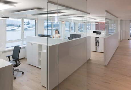 Modernes, helles Büro mit transparenten Glaswänden und weißen Trennelementen, die flexible Arbeitszonen definieren.