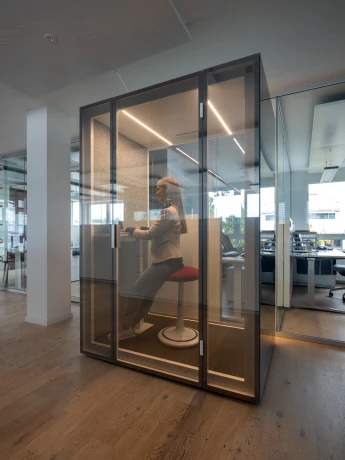 Frau in einem modern gestalteten, verglasten Raum innerhalb eines Büro- oder Ausstellungsbereichs. Minimalistische Einrichtung mit Schreibtisch.