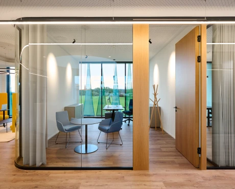 Moderner Büroeinrichtungsbereich mit Glasraumerweiterung. Ein Besprechungsbereich mit Tisch und Stühlen öffnet sich zu einer Fensterfront mit Außenblick.