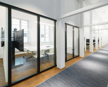 Moderne Büroszene mit großen Glaswänden, die einen hell beleuchteten Arbeitsbereich von einem breiten Flur trennen.